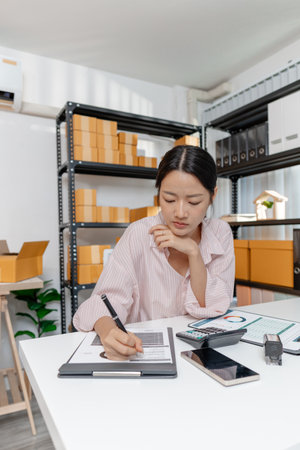 A young woman in a striped shirt works at her desk with documents, calculator, and smartphone,...