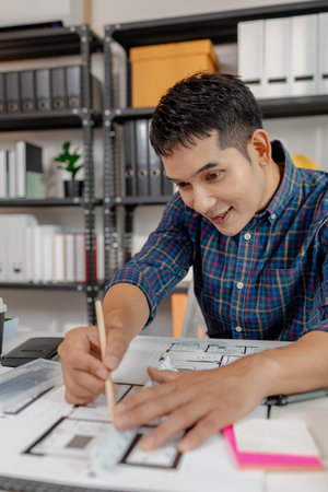 A young male architect is working with blueprints at his desk, using rulers and drawing tools to...