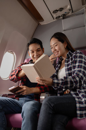 Asian couple sitting together on an airplane, reading a book and smiling while enjoying their journey. They share a pleasant moment during travel, creating memories and bonding as a couple.の写真素材