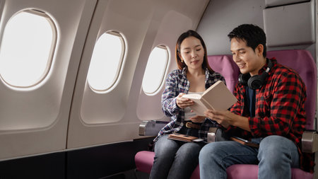 Asian couple sitting together on an airplane, reading a book and smiling while enjoying their...
