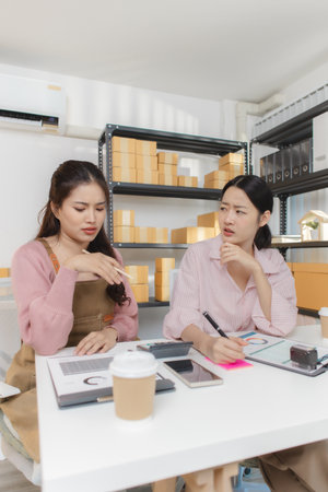 Two young women work together in a small business office, organizing packages, checking financial reports, and managing orders on digital devices, representing teamwork and entrepreneurship.の写真素材