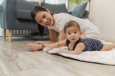 A baby enjoys tummy time on a soft mat while the mother stays close, encouraging him with a warm smile. This tender moment highlights early development, family care, and bonding at home.の写真素材