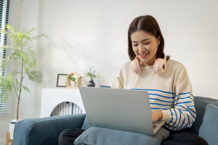 A young woman is sitting comfortably on a sofa using a laptop, smiling with focus and joy. She looks engaged in her work or online communication, creating a warm lifestyle concept.の写真素材