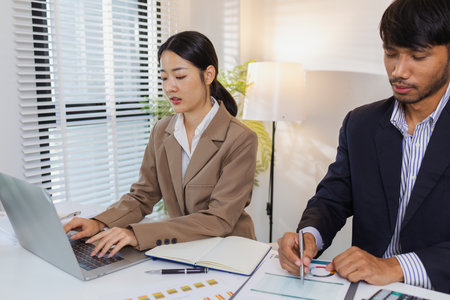 Two business professionals are discussing financial charts and reports in a modern office. They collaborate, share ideas, and analyze data together, highlighting teamwork and strategy.の写真素材