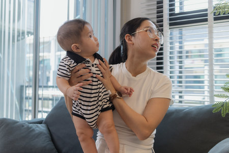 A young mother is holding her baby while spending quality time together indoors. She looks at her...