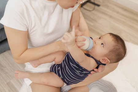 A mother is lovingly feeding her baby with a bottle of milk while holding the child in her arms. The...