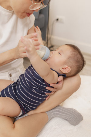 A mother is lovingly feeding her baby with a bottle of milk while holding the child in her arms. The...