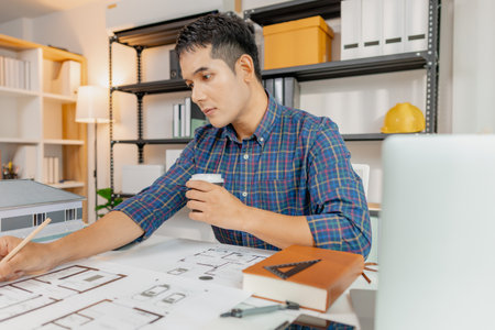 A young architect works on house design plans at his desk, focusing on drawings and scale models. The scene conveys creativity, dedication, and precision in modern architecture projects.の写真素材