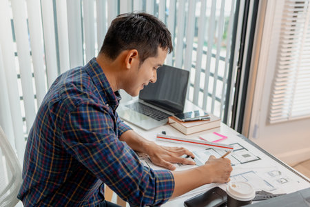A young male architect is working with blueprints at his desk, using rulers and drawing tools to refine details. He appears focused and engaged, balancing creativity with precision.の写真素材