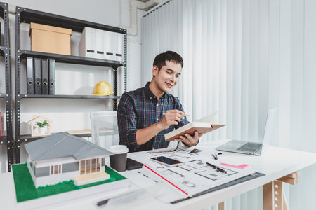 Young architect working in office with building models and floor plans, analyzing details and creating innovative design concepts, showing creativity, focus, and dedication in architecture projects.の写真素材