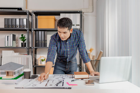 A young architect works with building plans and a house model in the office, reviewing blueprints, marking details, and analyzing design structures with focus and creativity.の写真素材