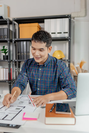 Confident young architect working at office desk, drawing house plans and discussing construction details on the phone, focusing on accuracy and creativity in architectural design and project planningの写真素材
