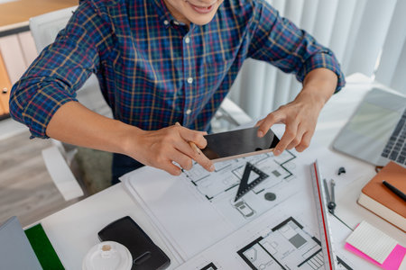 A young architect is multitasking at his desk, sketching blueprints, making phone calls, and reviewing notes. His workspace shows focus, productivity, and modern design work.の写真素材
