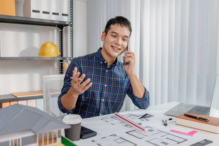 A young architect is multitasking at his desk, sketching blueprints, making phone calls, and...