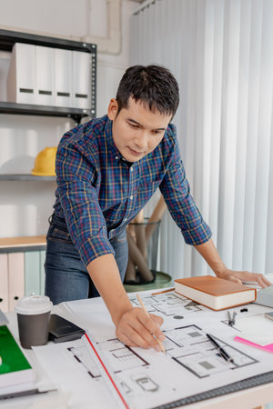 A young architect is multitasking at his desk, sketching blueprints, making phone calls, and reviewing notes. His workspace shows focus, productivity, and modern design work.の写真素材