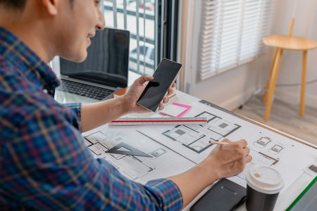 A young male architect is working with blueprints at his desk, using rulers and drawing tools to refine details. He appears focused and engaged, balancing creativity with precision.の写真素材