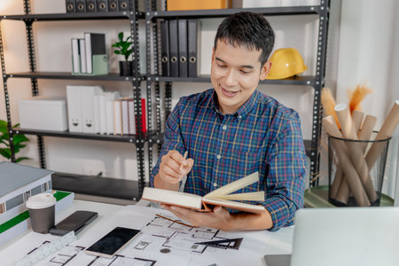 A young architect is multitasking at his desk, sketching blueprints, making phone calls, and reviewing notes. His workspace shows focus, productivity, and modern design work.の写真素材