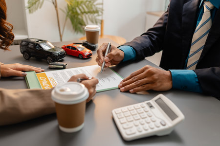 A businessperson and client are reviewing and signing a car sales contract at a desk, with small model cars, a coffee cup, and documents around them, symbolizing vehicle purchase negotiation.の写真素材