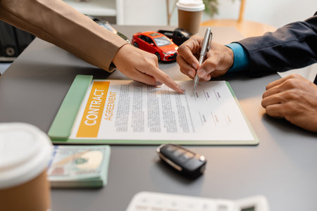 A businessperson and client are reviewing and signing a car sales contract at a desk, with small model cars, a coffee cup, and documents around them, symbolizing vehicle purchase negotiation.の写真素材