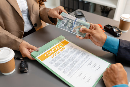 A businessperson and client are reviewing and signing a car sales contract at a desk, with small model cars, a coffee cup, and documents around them, symbolizing vehicle purchase negotiation.の写真素材