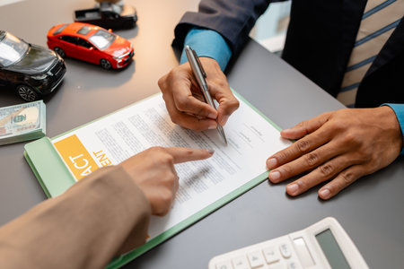 A businessperson and client are reviewing and signing a car sales contract at a desk, with small model cars, a coffee cup, and documents around them, symbolizing vehicle purchase negotiation.の写真素材