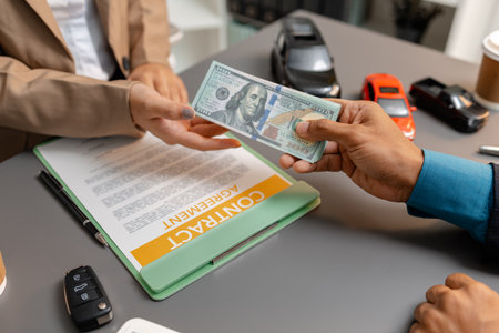 A businessperson and client are reviewing and signing a car sales contract at a desk, with small model cars, a coffee cup, and documents around them, symbolizing vehicle purchase negotiation.の写真素材