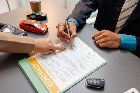 A businessperson and client are reviewing and signing a car sales contract at a desk, with small...
