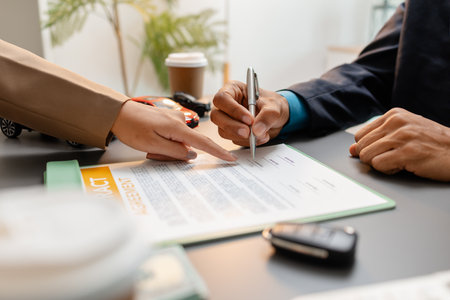 A businessperson and client are reviewing and signing a car sales contract at a desk, with small model cars, a coffee cup, and documents around them, symbolizing vehicle purchase negotiation.の写真素材