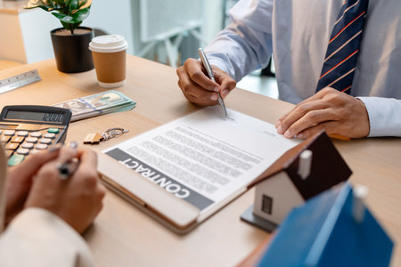 A real estate agent and a client are discussing and pointing at a housing contract document during a property purchase meeting, showing agreement details and financial negotiation about home ownershipの写真素材