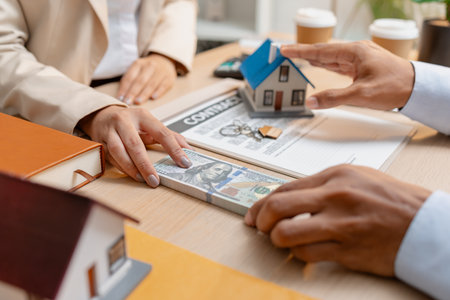 A real estate agent presents a property sale contract to a client, discussing agreement details with a model house on the document, symbolizing home buying, selling, and real estate investment.の写真素材