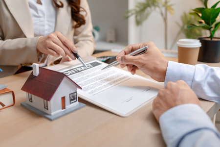 A real estate agent and a client are discussing and pointing at a housing contract document during a property purchase meeting, showing agreement details and financial negotiation about home ownershipの写真素材