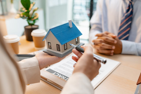 A real estate agent presents a property sale contract to a client, discussing agreement details with a model house on the document, symbolizing home buying, selling, and real estate investment.の写真素材