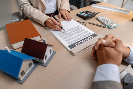A real estate agent and client discussing property sale details at the office table, reviewing and signing home purchase contracts with miniature house models and money on the desk.の写真素材
