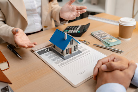 A real estate agent presents a property sale contract to a client, discussing agreement details with a model house on the document, symbolizing home buying, selling, and real estate investment.の写真素材