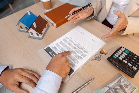 A real estate agent and client discussing property sale details at the office table, reviewing and signing home purchase contracts with miniature house models and money on the desk.の写真素材