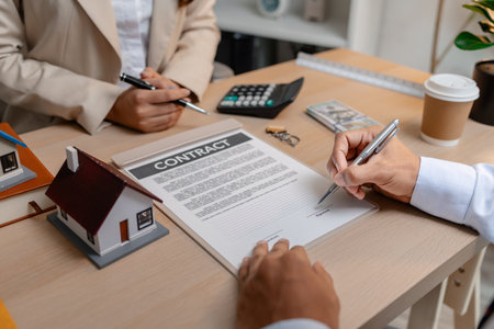 A real estate agent and a client are discussing and pointing at a housing contract document during a property purchase meeting, showing agreement details and financial negotiation about home ownershipの写真素材