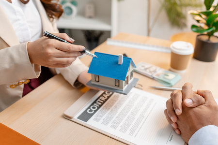 A real estate agent presents a property sale contract to a client, discussing agreement details with a model house on the document, symbolizing home buying, selling, and real estate investment.の写真素材