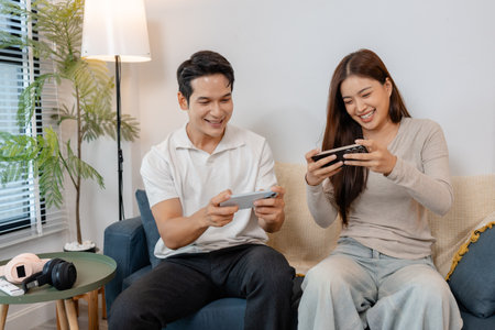A cheerful Asian couple sitting together on a sofa, playing mobile games and laughing happily in a cozy living room. They enjoy their leisure time and friendly competition with joy.の写真素材