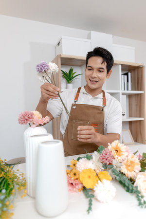 A smiling man wearing an apron arranges colorful flowers in a vase on a table in a cozy room. He appears focused and content while creating a beautiful floral arrangement as part of his work.の写真素材