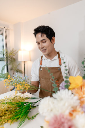 A smiling man wearing an apron arranges colorful flowers in a vase on a table in a cozy room. He appears focused and content while creating a beautiful floral arrangement as part of his work.の写真素材