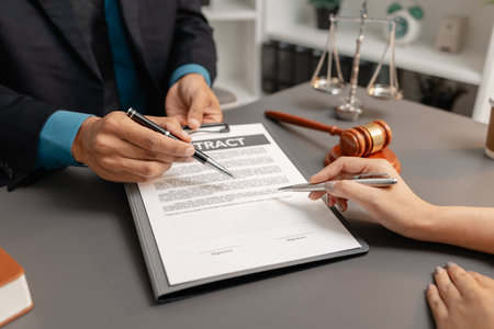 A male lawyer in a suit discusses contract details with a female client, pointing to the signature...