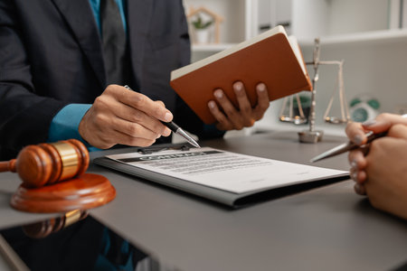 A male lawyer in a suit discusses contract details with a female client, pointing to the signature area on a legal document. The office desk features a gavel symbolizing law and justice.の写真素材