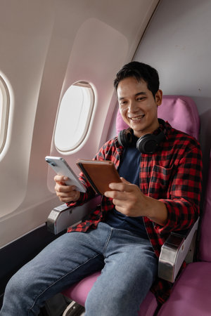 A young man sits on an airplane, enjoying his flight while checking his smartphone and passport. He...