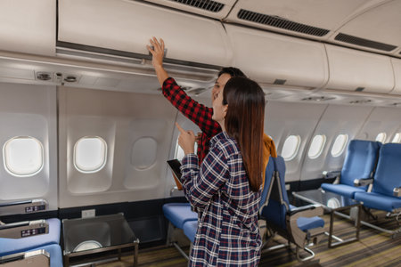 A young couple on an airplane. The man places luggage in the overhead bin while the woman smiles, both appearing excited.の写真素材