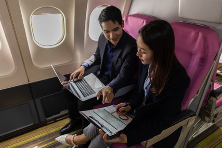 A business man and woman in formal suits working together on a flight. They are discussing documents and using laptops, showing teamwork and productivity while traveling on an airplane.の写真素材