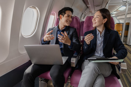 A business man and woman in formal suits working together on a flight. They are discussing documents and using laptops, showing teamwork and productivity while traveling on an airplane.の写真素材