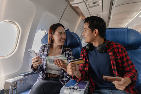 A happy couple sitting together on an airplane, holding cash, credit cards, and smartphones. They appear to be managing their travel expenses or planning their spending during the flight.の写真素材