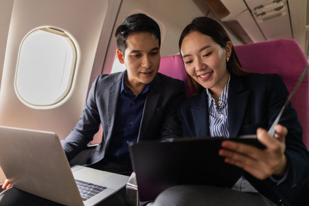A business man and woman in formal suits working together on a flight. They are discussing documents and using laptops, showing teamwork and productivity while traveling on an airplane.の写真素材