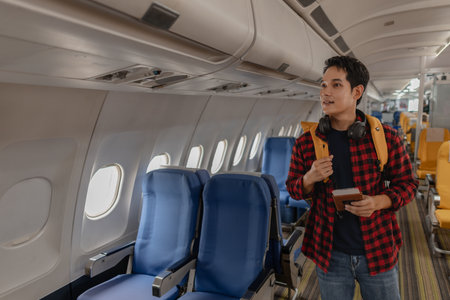 Asian man boarding an airplane and searching for his seat with a passport and boarding pass in hand. He looks happy and excited, carrying a backpack and wearing headphones around his neck.の写真素材