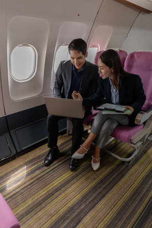 A business man and woman in formal suits working together on a flight. They are discussing documents and using laptops, showing teamwork and productivity while traveling on an airplane.の写真素材